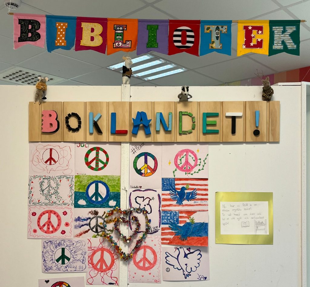 Entrén till skolbiblioteket "Boklandet" med en färgglad flaggbanderoll med texten "Bibliotek" och en träskylt med bokstäver, omgiven av elevers teckningar med fredstecken och en informationslapp om nya engelska böcker.