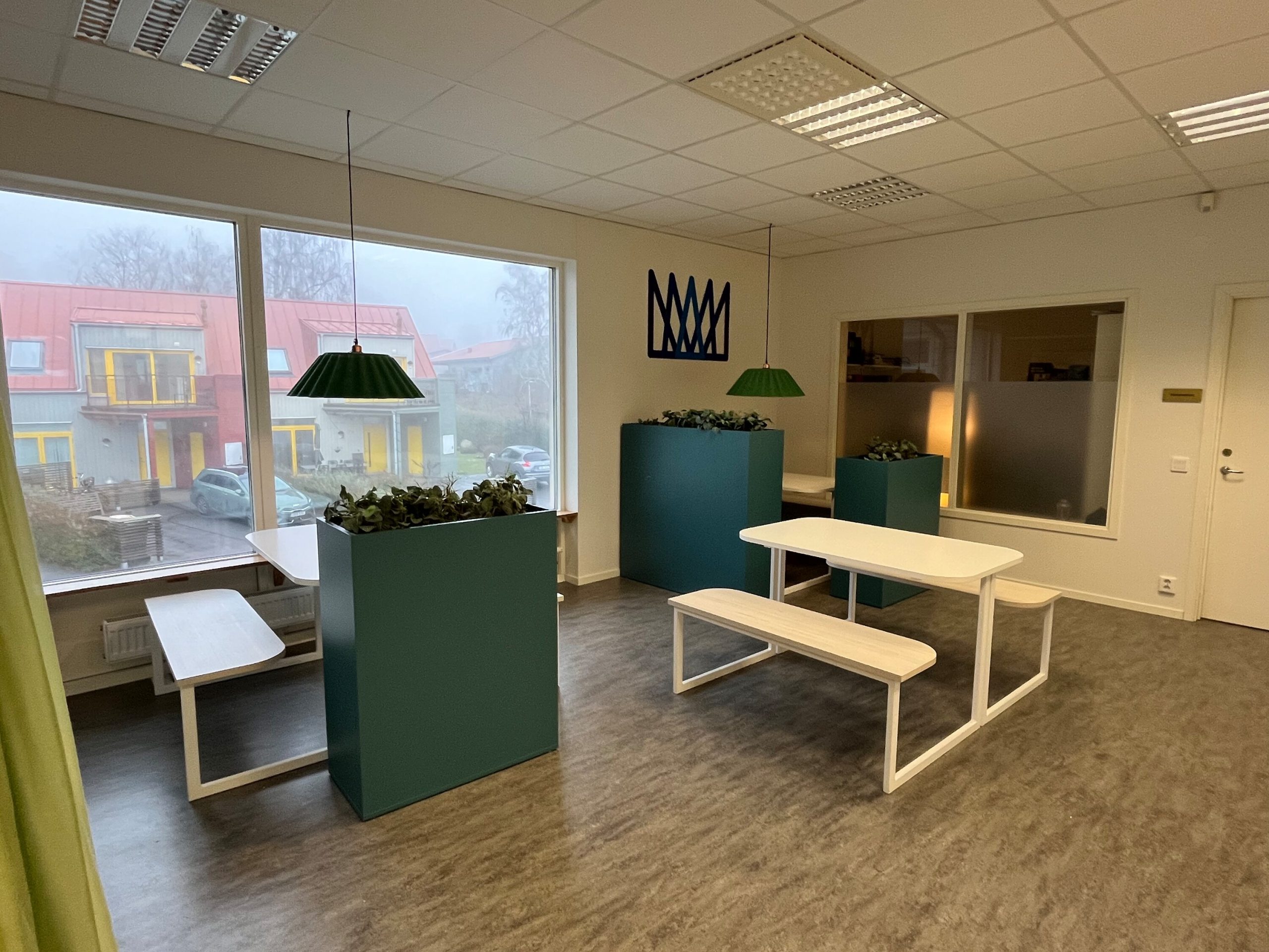 Ett ljust gemensamt utrymme med vita bänkbord, gröna växtavdelare och en grön hängande taklampa, med stora fönster ut mot omgivande bebyggelse.
