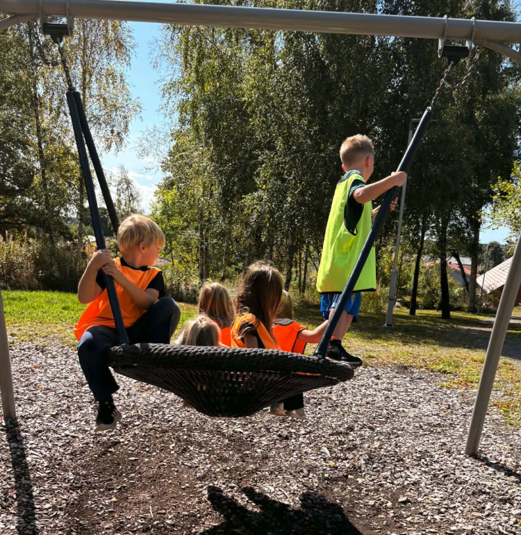 Flera barn i reflexvästar sitter tillsammans i en stor nätgunga på en lekplats en solig dag, omgiven av träd och grönska.