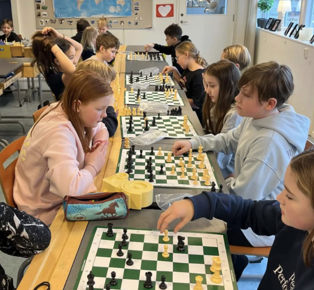 Elever sitter parvis och spelar schack vid flera bord i ett klassrum med en världskarta på väggen, koncentrerade på sina schackspel.